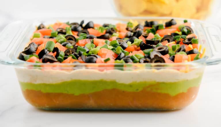 Layered Taco Dip in Glass Dish with Bowl of Chips in the Back