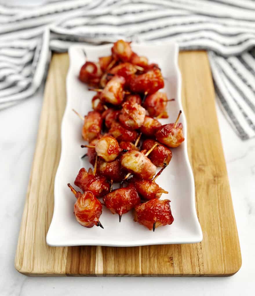 Bacon Wrapped Water Chestnuts on White Serving Platter on Wooden Cutting Board with Kitchen Towel in the Background