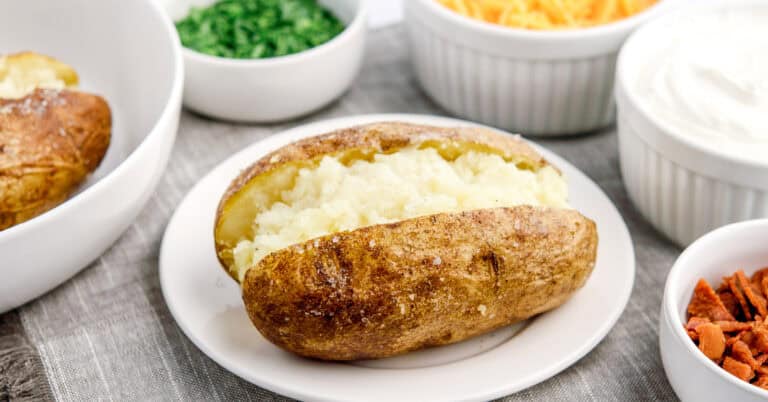 Baked Potato on White Plate with Bowls of Toppings Behind