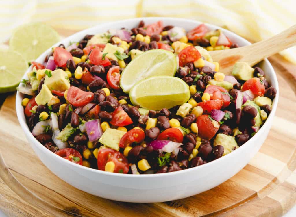 Black Bean Salad With Corn and Avocado in Serving Bowl With Limes and Wooden Spoon