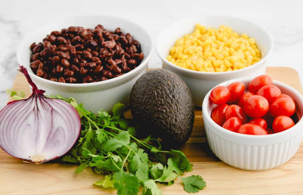Black Beans, Corn, and Cherry Tomatoes in White Bowls on a Cutting Board with Half a Red Onion, Cilantro, and Avocado