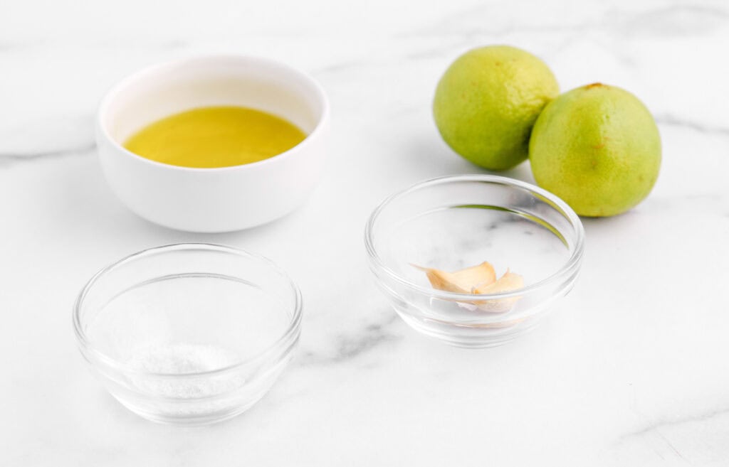 Olive Oil, Garlic Cloves, and Salt in Bowls and Fresh Limes on White Marble Table