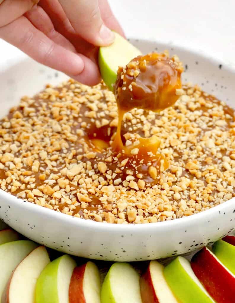 Dipping Green Apple in Caramel Apple Dip in Bowl with Apple Slices on Platter