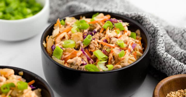Egg Roll in a Bowl in a Black Bowl with Chopped Green Onions in a White Bowl in the Background with a Kitchen Towel