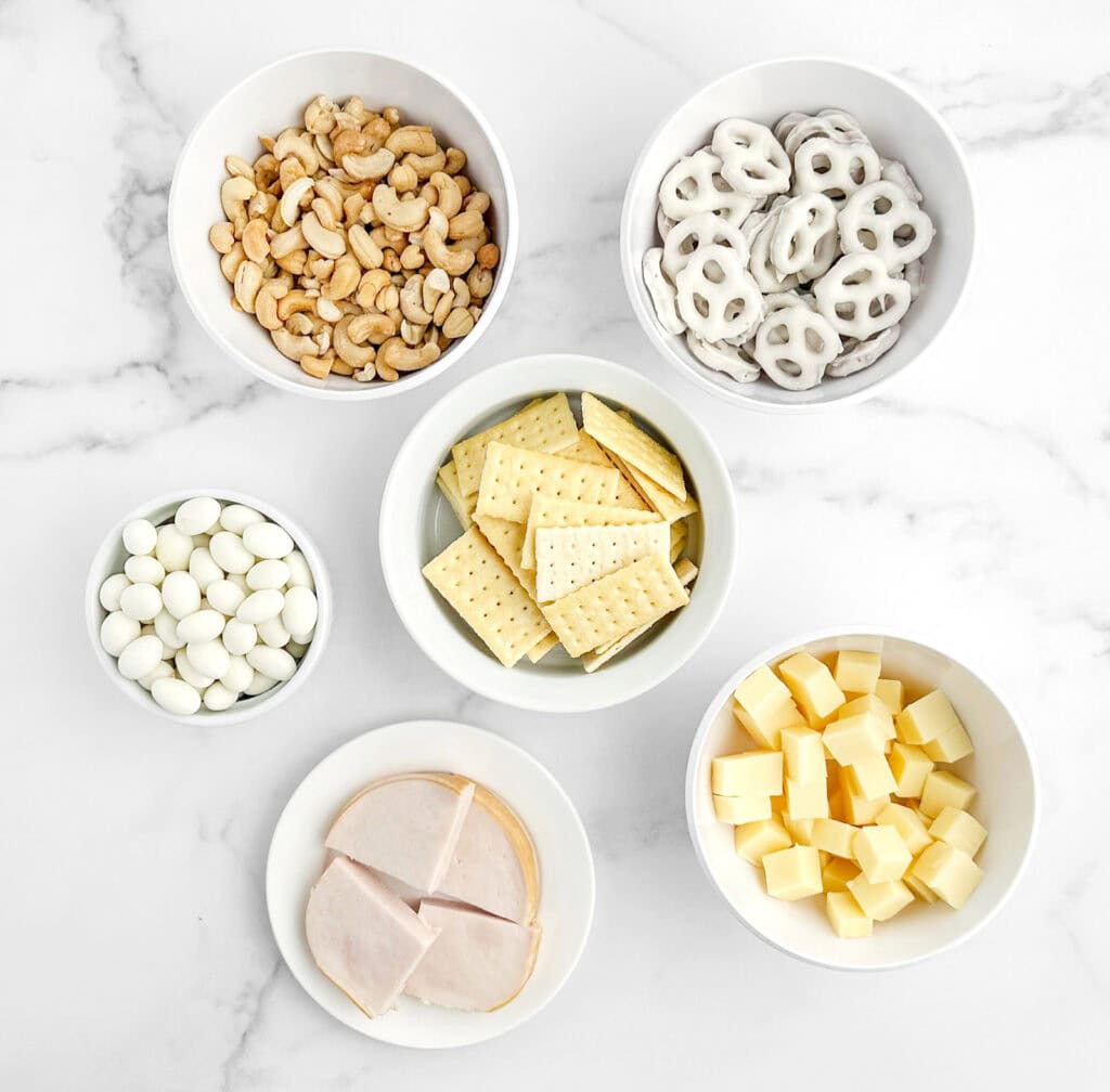White Ingredients in White Bowls on Marble Surface (White Chocolate Covered Pretzels, Yogurt Almonds,  Cheeses, Turkey Slices)