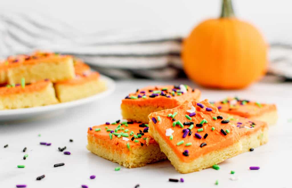 Halloween Frosted Sugar Cookie Bars in Pile with Plate of Bars in Background with Pumpkin