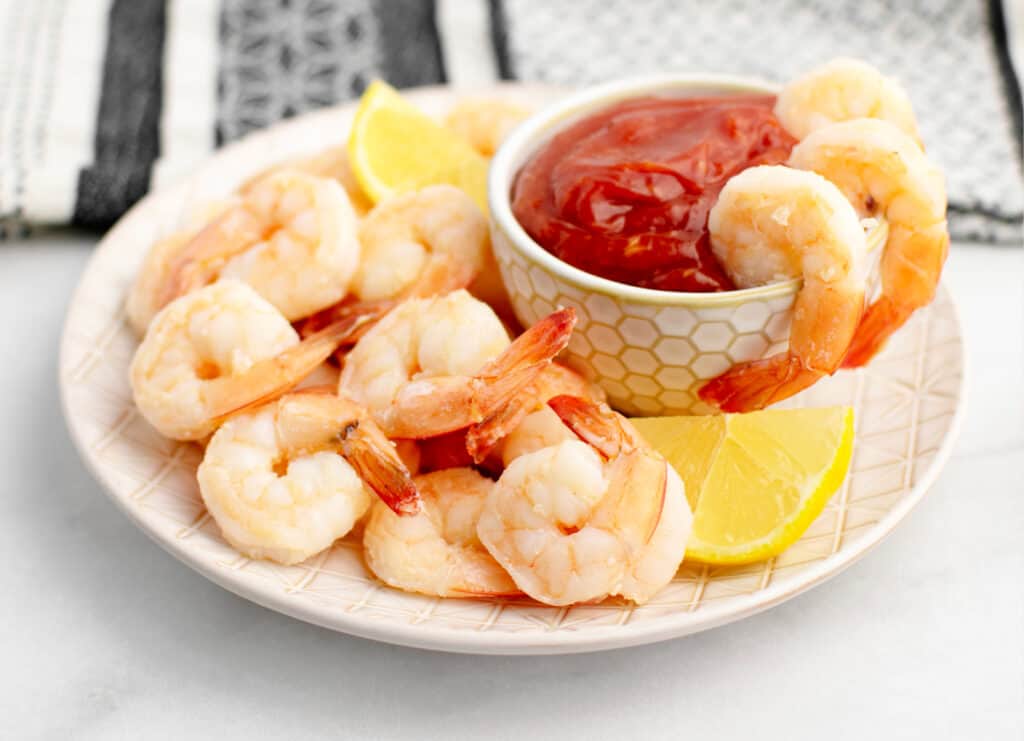 Shrimp Cocktail with Cocktail Sauce in Bowl and Shrimp Pieces on the Side on a Plate with More Shrimp and Lemons