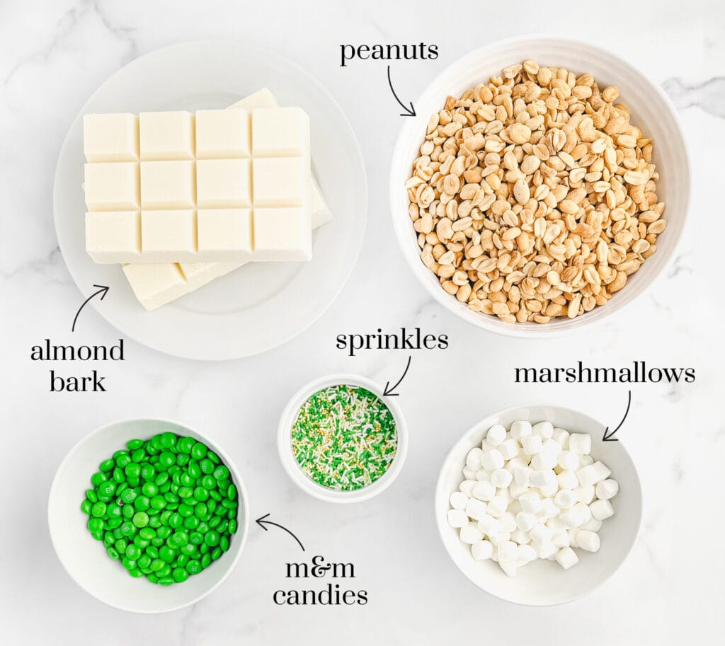 Almond Bark, Peanuts, Green M&Ms, Sprinkles, and Mini Marshmallows in Bowls on Marble Surface with Labels